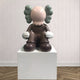 The Hazelnut Brown Iconify Thinker Sitting Sculpture by Giant Sculptures stands at 92cm, featuring a round head, X-shaped eyes, and large gloves. Its distinctive ears and hazelnut tones are showcased on a white pedestal in a room with wood flooring, exuding modern artistry.