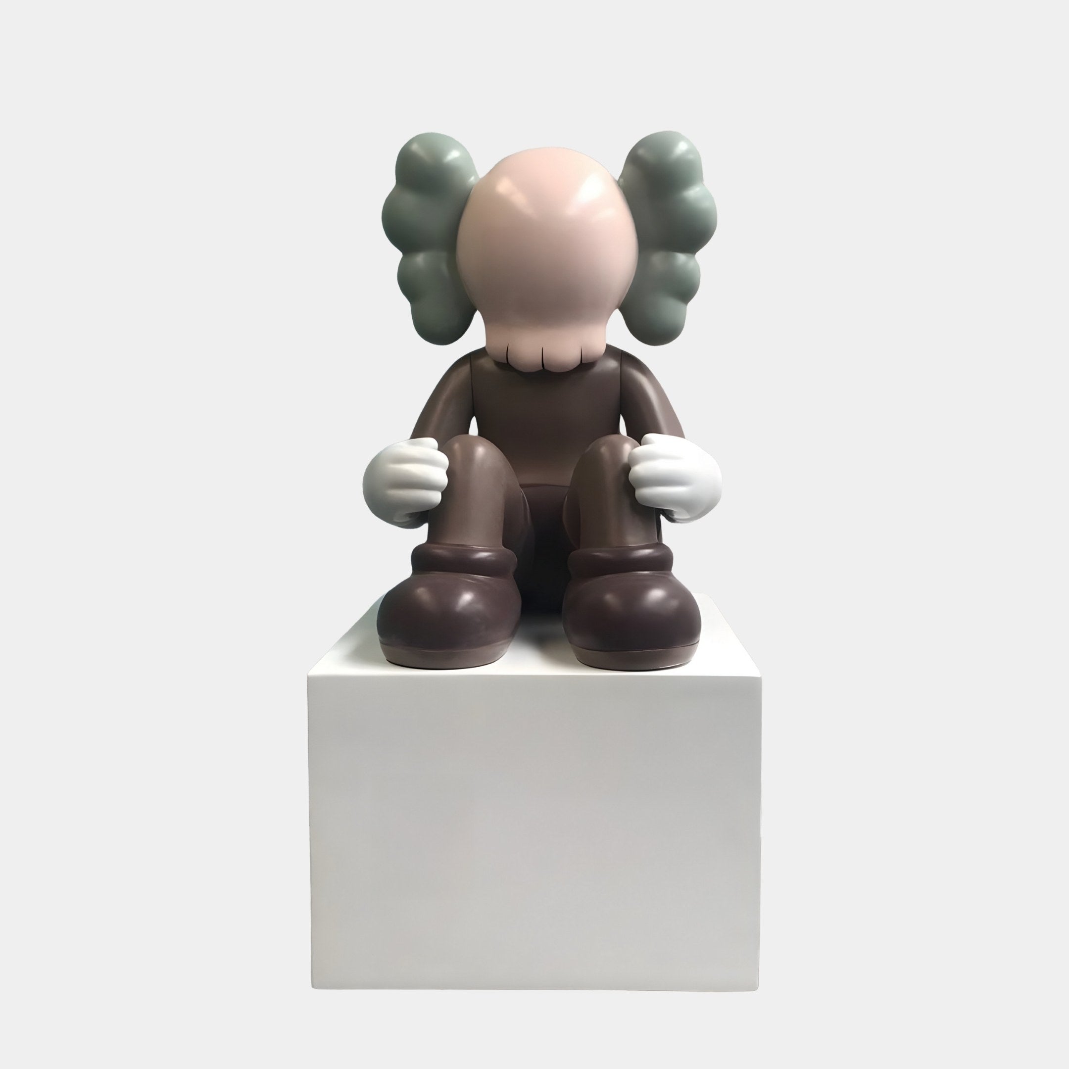 The Hazelnut Brown Iconify Thinker Sitting Sculpture by Giant Sculptures is a modern art piece featuring a cartoonish figure with rounded gray ears, gloved hands, a faceless hazelnut-brown face, seated thoughtfully on a white block and dressed in dark clothing.