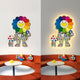 The Clovelle Floral Bliss UV Wood Carving LED Wall Art by Giant Sculptures, featuring two colorful smiling characters, adorns a white wall above an orange couch. A round table with toy figures sits in front, with the left character smaller and the right one larger and illuminated.