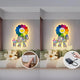 Two images depict the Clovelle Floral Bliss UV Wood Carving LED Wall Art by Giant Sculptures, showcasing colorful floral artwork and smiley faces on a bedroom wall. One image shows a finger at a light switch; the other features a remote with a black control box. A bed with round pillows and a green headboard is in the background.