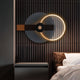 In a modern bedroom, the Eclipse I Charcoal Bronze Acrylic 3D LED Wall Art by Giant Sculptures decorates a wood-slatted backdrop. White bedding and a brown pillow on the bed are paired with a minimalist nightstand, enhancing the rooms neutral tones.