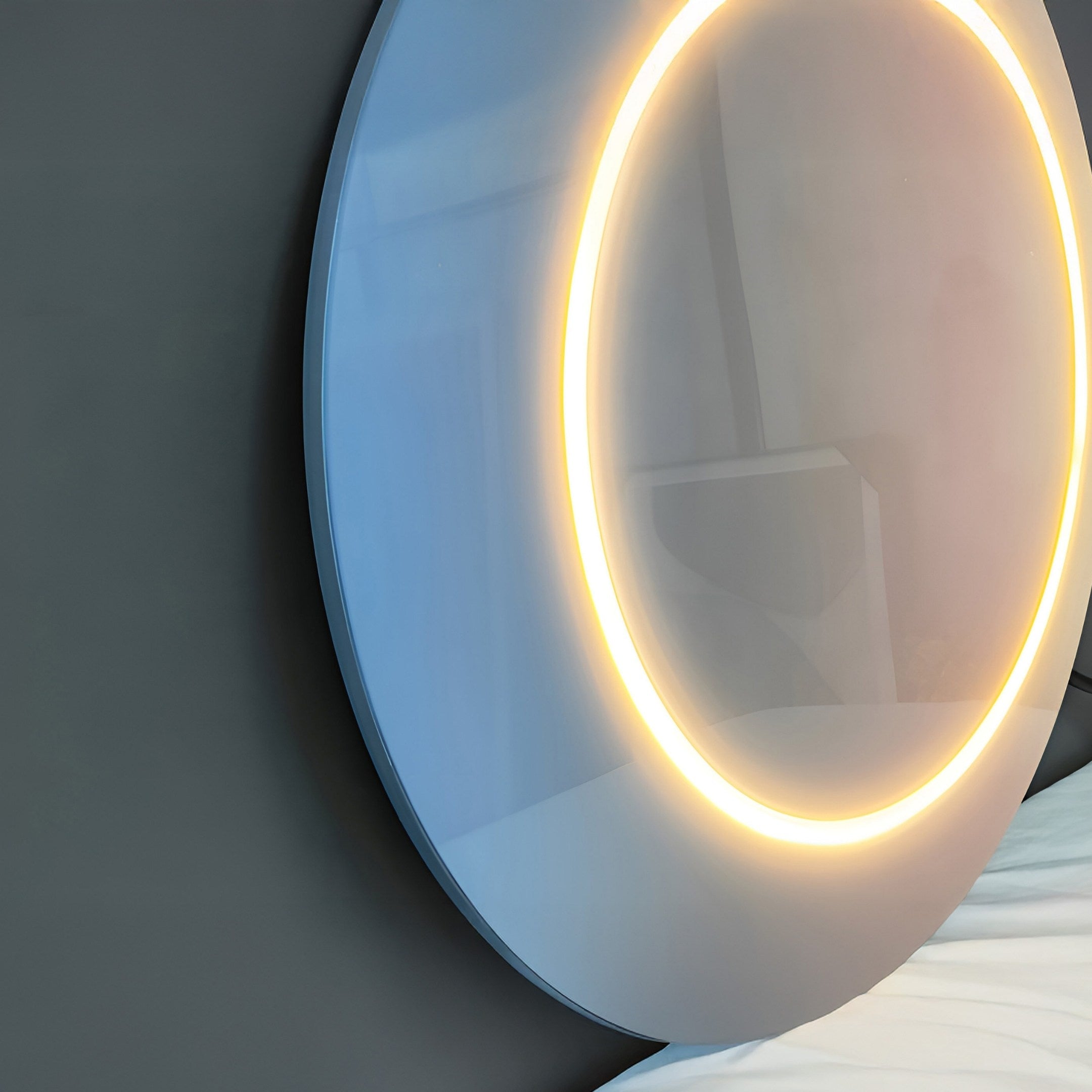 The Aurora Blush Mist Crystal Porcelain LED Wall Art by Giant Sculptures, resembling a glowing round mirror, is mounted above a bed on a dark wall. Its reflection suggests the interiors modern and minimalist vibe.