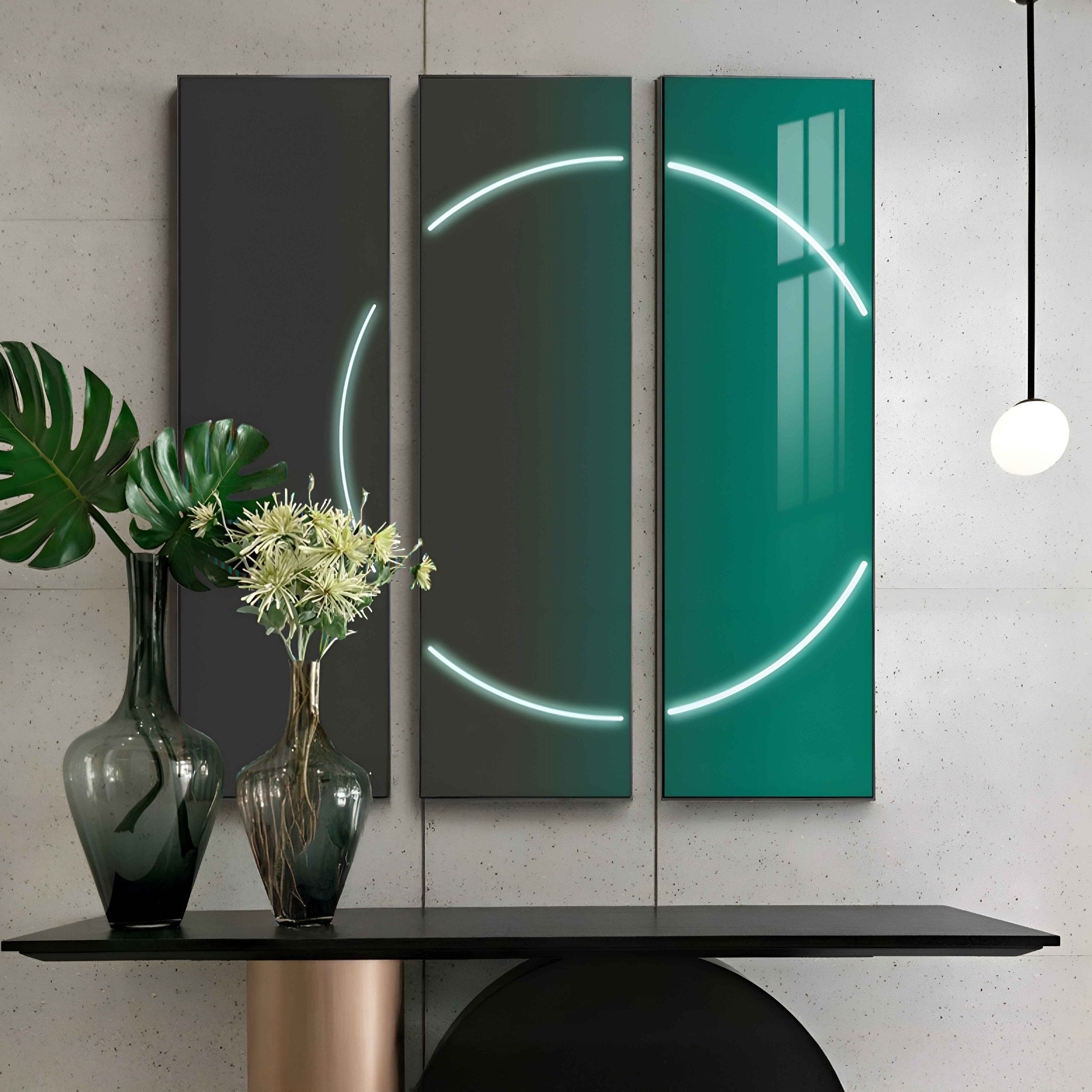 The Helios Emerald Crystal Porcelain LED Wall Art from Giant Sculptures showcases a continuous neon circle across three vertical panels in green gradient and dark grey tones. A modern vase with tropical leaves and white flowers sits on a minimalist table beneath, epitomizing stylish modern interiors.