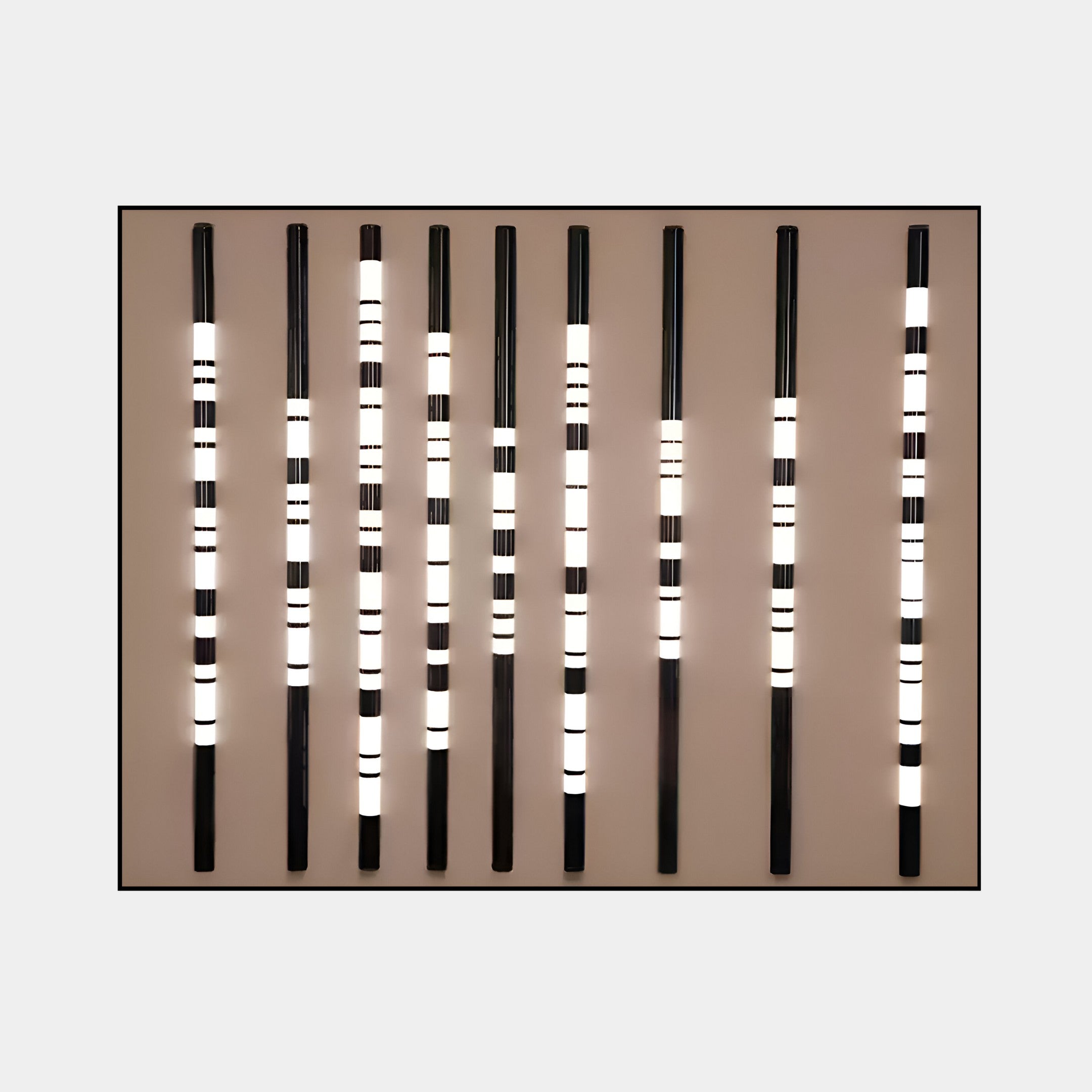 The Strata Taupe LED Wall Art from Giant Sculptures features nine vertical black rods with lit white sections on a taupe background, creating a minimalist light-and-dark pattern ideal for modern homes.