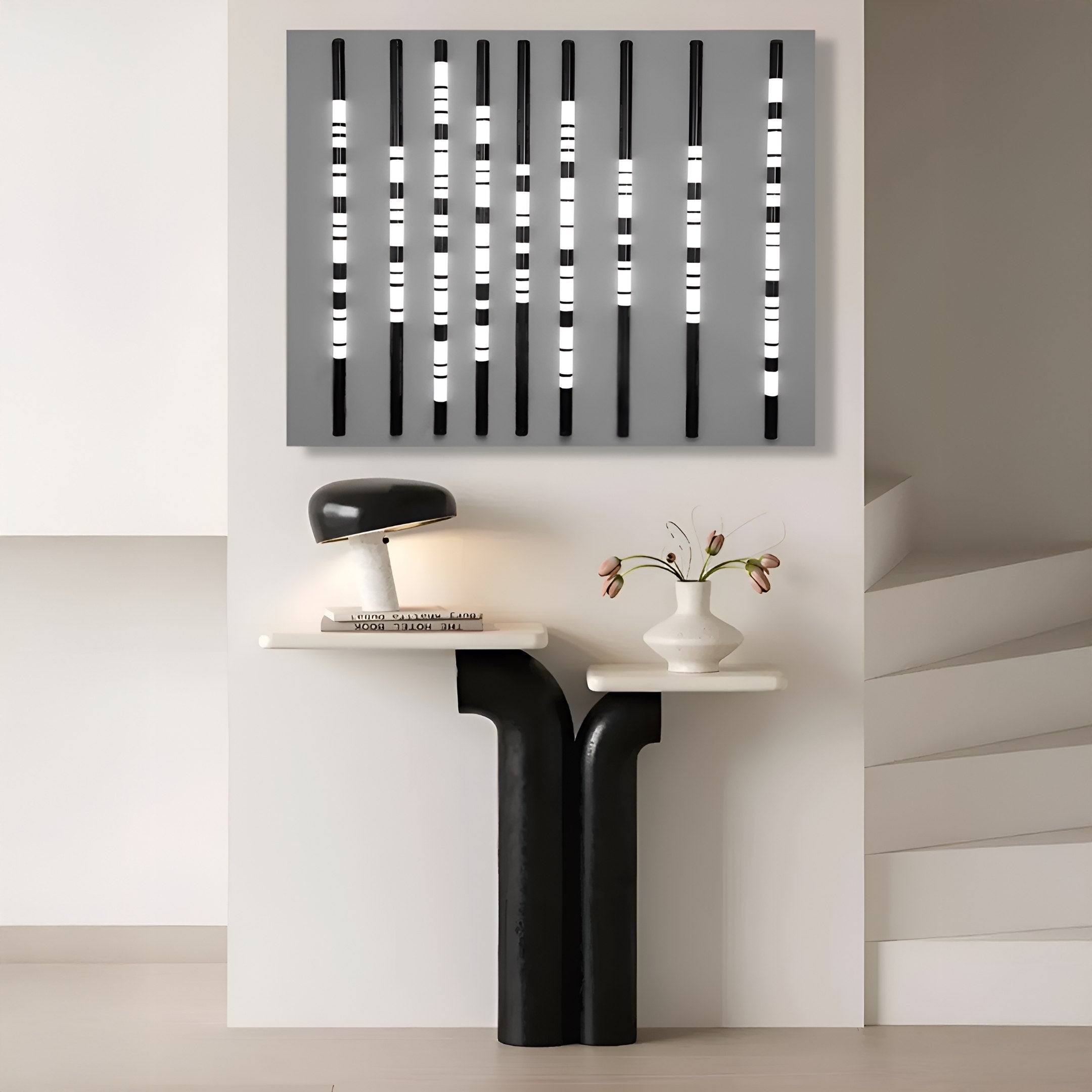 A modern interior features a sleek black and white table with asymmetric legs, topped with a lamp, books, and flowers. Above it hangs Giant Sculptures Strata Silver LED Wall Art, casting an elegant glow through its integrated LEDs amidst vertical black and white bars.