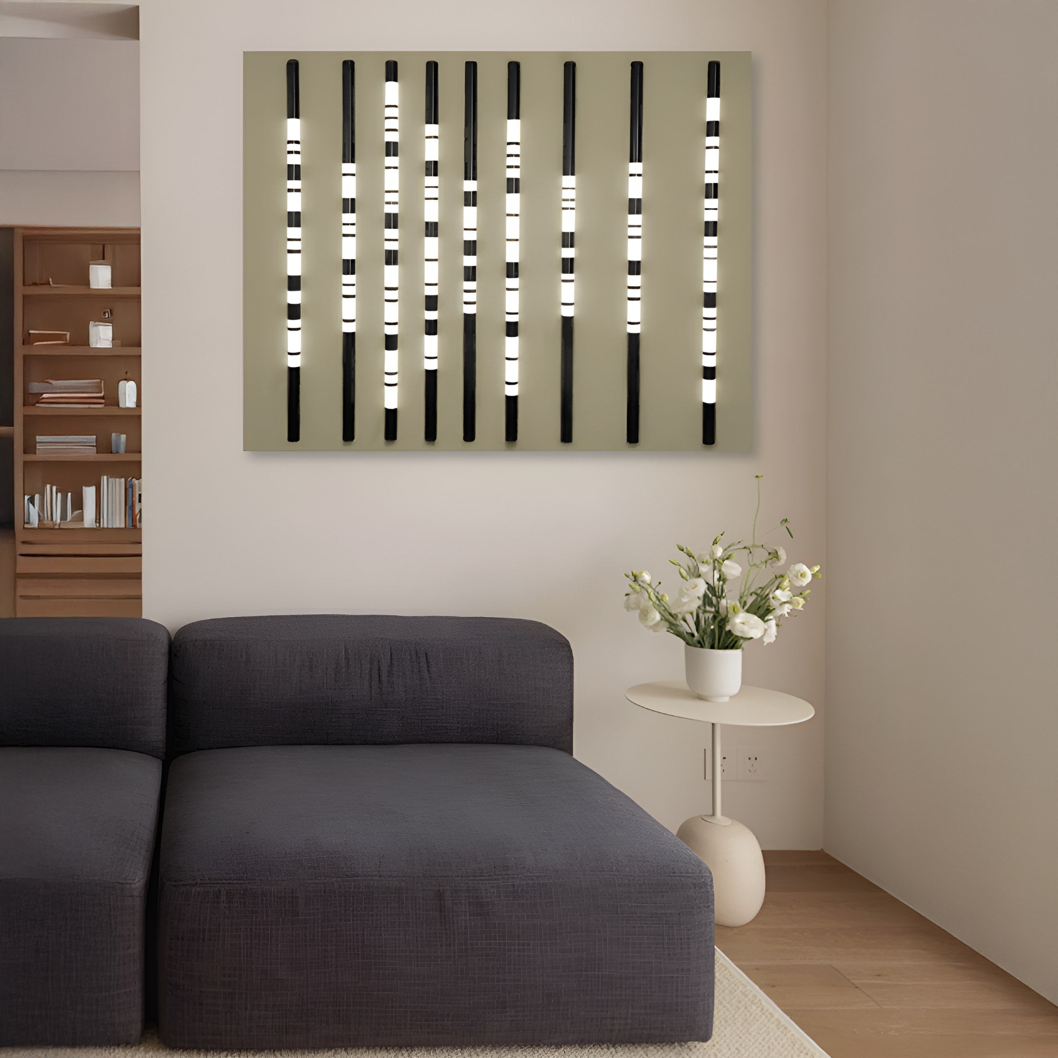 A modern living room features a slate-gray sectional with a white side table and flowers. Strata Slate LED Wall Art from Giant Sculptures, boasting vertical black and white stripes, adds flair, while shelves on the left display books and items.