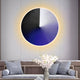 A modern living room showcases the Orbit Violet Hand-Painted Canvas 3D LED Wall Art by Giant Sculptures, featuring black, white, and blue segments with a glowing halo. Below, a sofa with gray pillows and a table adorned with a white floral arrangement complete the decor.