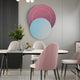 A modern dining room boasts an elegant oval table surrounded by four pink and white upholstered chairs. The gray wall features the Lunar Blush Sky Crystal Porcelain 3D White LED Wall Art by Giant Sculptures, with purple and blue circles. A vase with foliage and a decorative book add charm to the table.