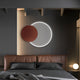 A modern bedroom with a leather headboard and neatly made bed features Giant Sculptures Soluna Copper Acrylic 3D LED Wall Art, displaying a circular LED light and smaller red disc. A small spherical lamp on a bedside table adds extra lighting.