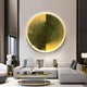 In a modern living room, the Astral Gold Crystal Porcelain LED Wall Art from Giant Sculptures showcases a large circular abstract piece. A gray sectional sofa complements a black glass coffee table with an aluminum frame, creating neutral decor and warm ambiance.