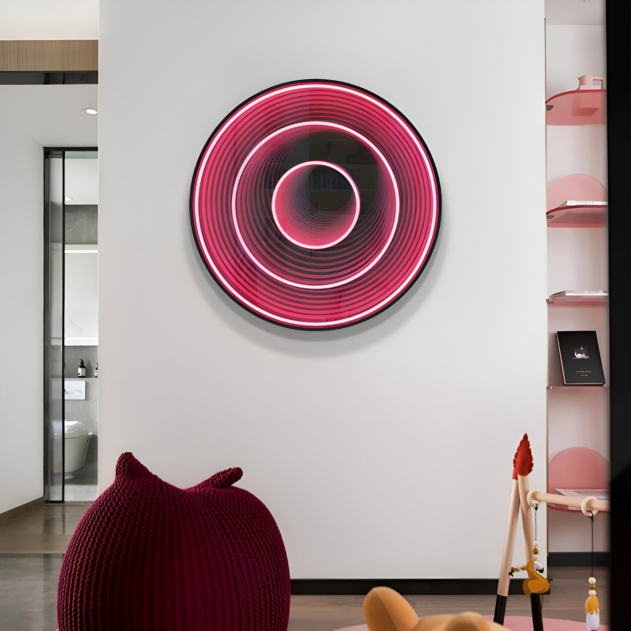 A modern interior features the Reflex Crimson Mirror 3D LED Wall Art by Giant Sculptures on a white wall, its rings glowing pink and purple. A red knitted pouf sits in front, with a reflective surface nearby and a glass partition with a door on the left. A pink shelf is positioned on the right.