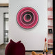 A modern interior features the Reflex Crimson Mirror 3D LED Wall Art by Giant Sculptures on a white wall, its rings glowing pink and purple. A red knitted pouf sits in front, with a reflective surface nearby and a glass partition with a door on the left. A pink shelf is positioned on the right.