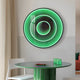 The Reflex Emerald Mirror 3D LED Wall Art by Giant Sculptures is mounted above a round table holding a green rectangular object and a small plate with red items. Nearby, theres a white chair, and a beige curtain is partially visible.