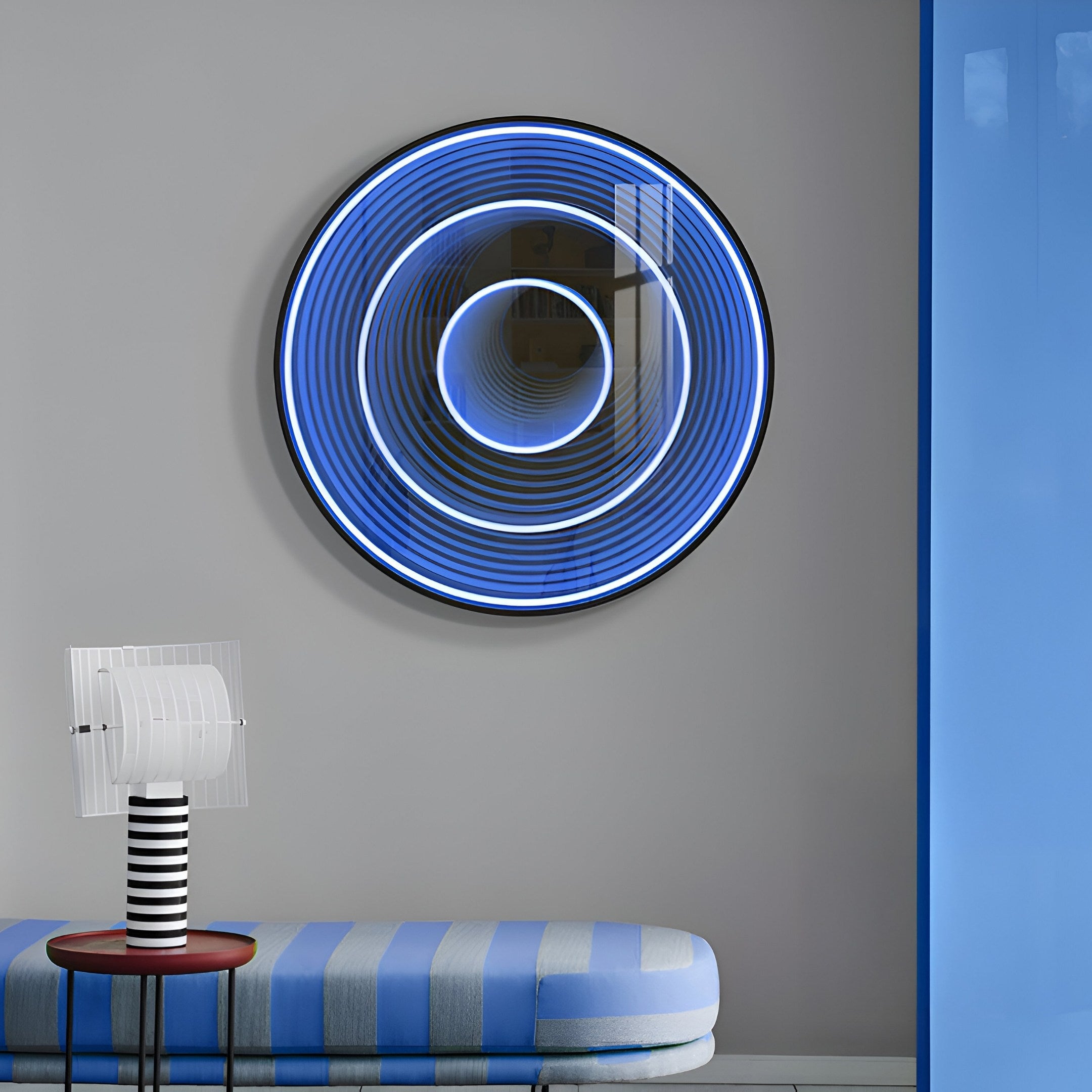 A modern interior features the Giant Sculptures Reflex Sapphire Mirror 3D LED Wall Art with blue and white concentric circles. Below, a striped bench with plush cushions sits next to a lamp with a transparent shade on a striped base, complemented by glimpses of the sapphire mirror partition.