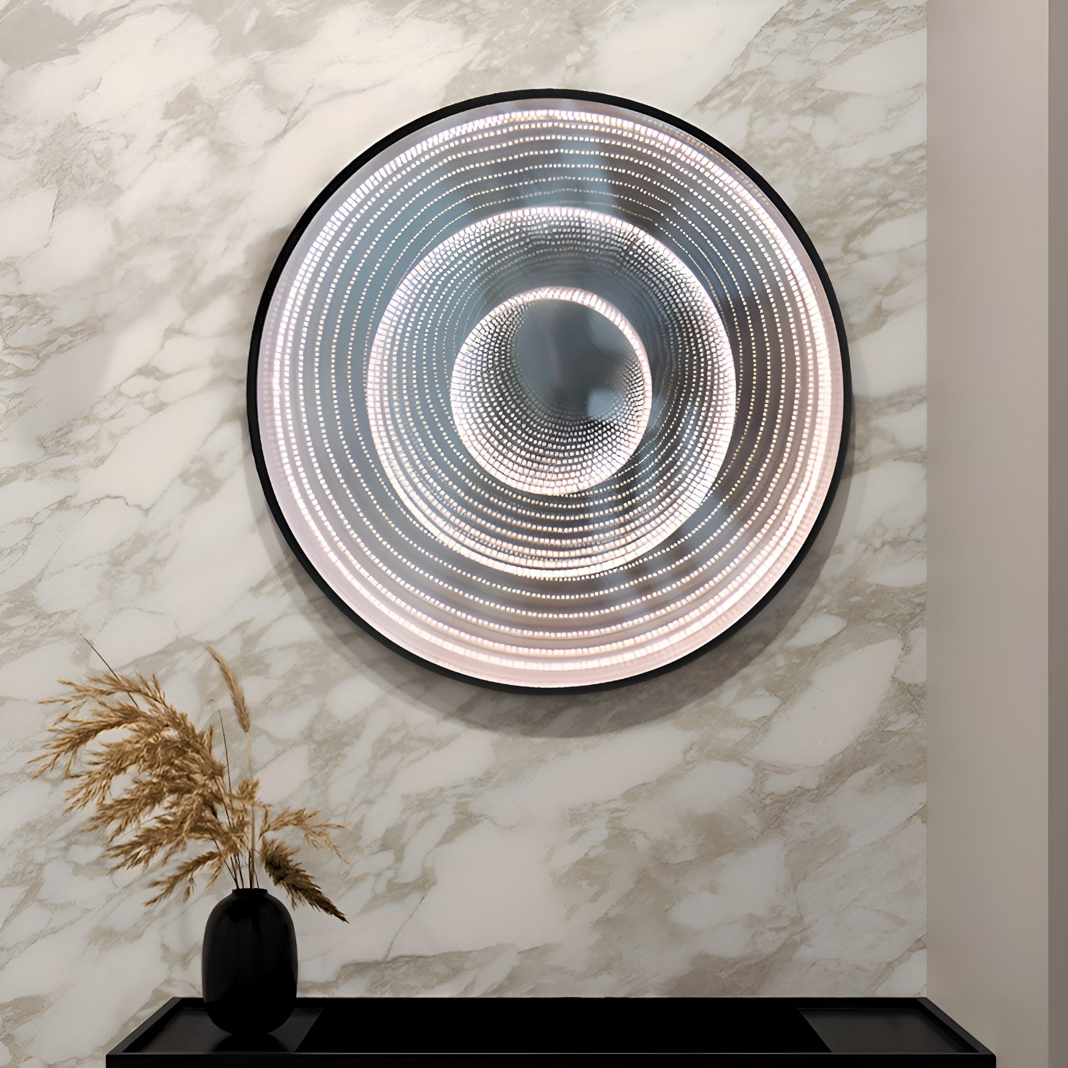 The Reflex Silver Mirror LED 3D Wall Art by Giant Sculptures, featuring a spiral pattern optical illusion, elegantly hangs on a marble-patterned wall. Below it, a black vase filled with dried grass completes the sophisticated home decor.