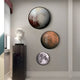 A modern room showcases a Pluto-inspired design featuring a bust on a black marble pedestal. Illuminated by natural light through window blinds, the Galaxia Pluto Crystal Porcelain LED Wall Art by Giant Sculptures graces the wall, evoking distant planets with its varied surfaces and colors.