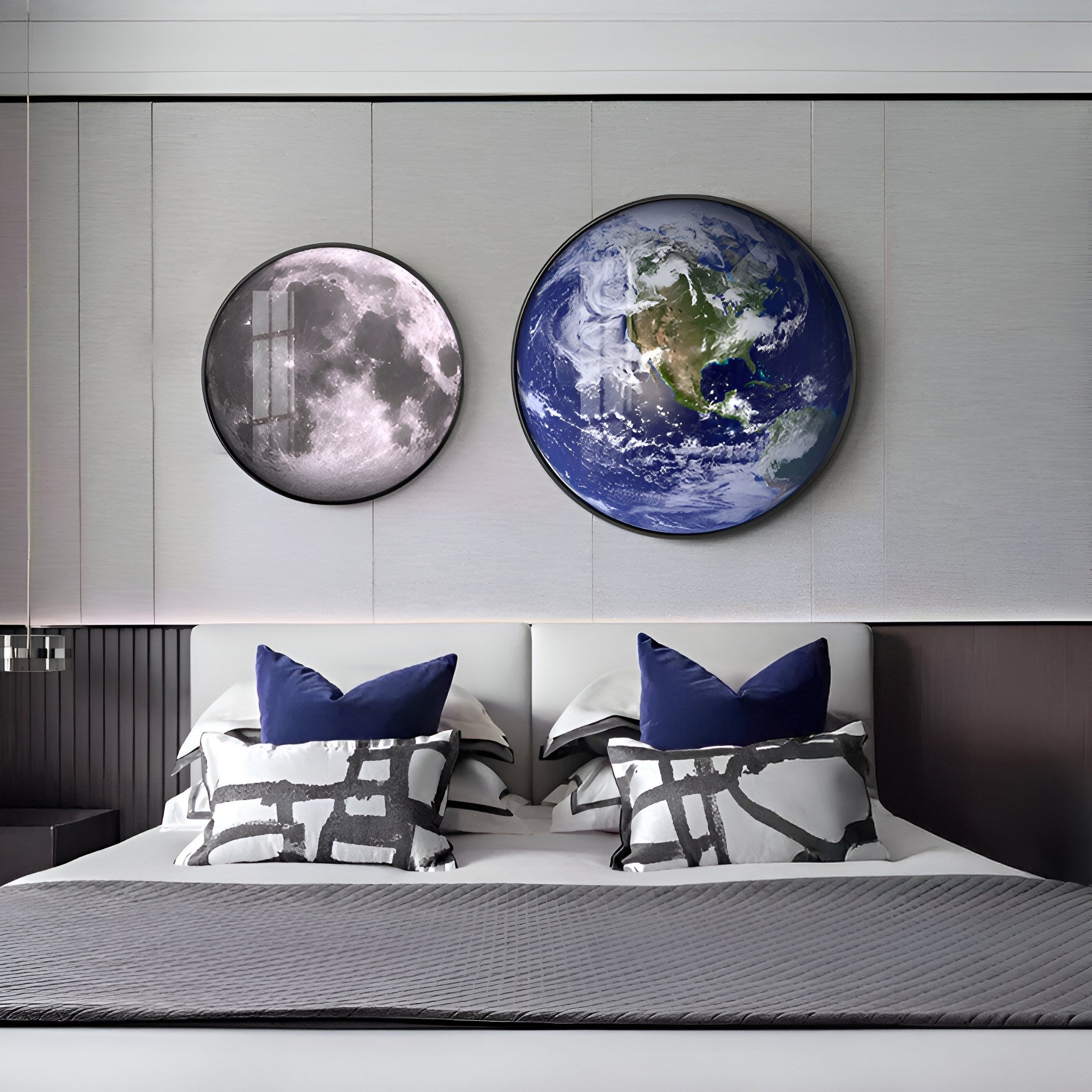 A modern bedroom showcases a bed with gray and white patterned bedding, blue pillows, and two round framed art pieces. One features the Moon, while the other is the Galaxia Earth Crystal Porcelain LED Wall Art by Giant Sculptures—all elegantly framed in brushed aluminum.