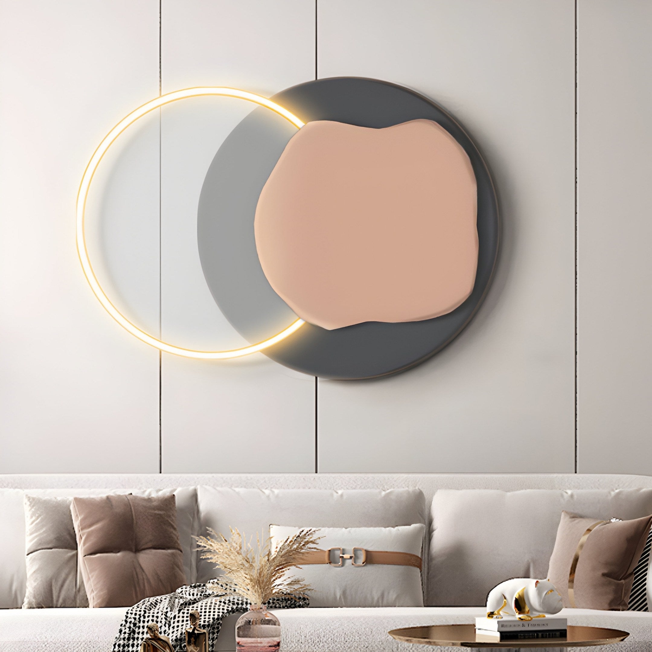 Embracing modern minimalism, the living room highlights the Arcadia Graphite Blush Leather 3D LED Wall Art by Giant Sculptures, featuring overlapping circles with a warm glow. A beige sofa adorned with throw pillows pairs seamlessly with a small table holding a decorative vase filled with dried grasses.