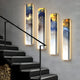 The Chroma White Ethereal Glow LED Wall Art Canvas by Giant Sculptures, featuring vertical abstracts in blue, gold, and white, is mounted on a light gray wall with backlit warmth. A sleek black staircase with stacked firewood ascends on the left in this modern interior.