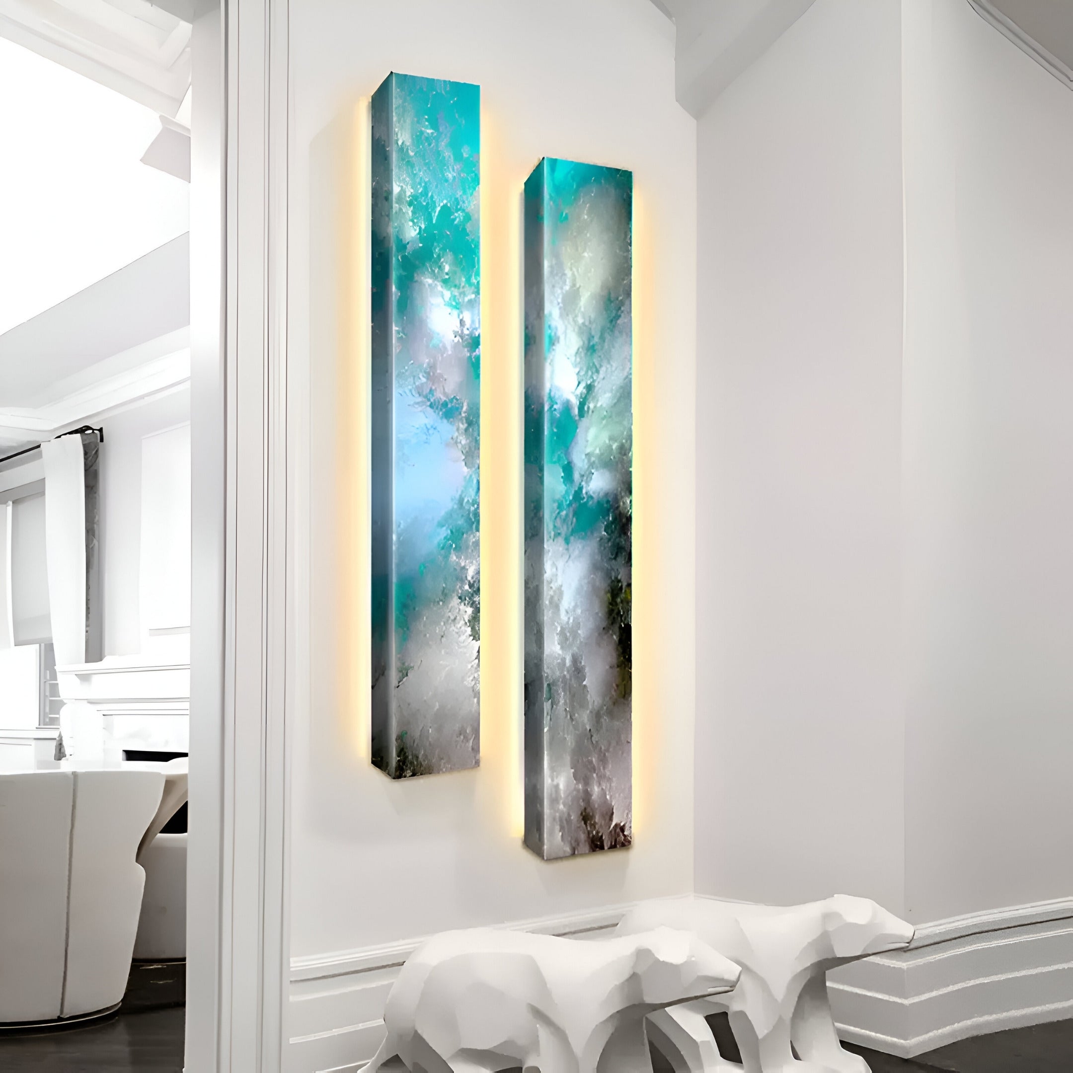 Two Chroma Sky Turquoise Mist LED Wall Art canvases by Giant Sculptures, showcasing abstract green, blue, and turquoise tones, are mounted on a white wall with soft backlighting. Below them sits a contemporary white sculpture enhancing the allure of modern interiors.