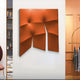 The Contours Bold Orange Elastic Fabric 3D Wooden Wall Art by Giant Sculptures, featuring bold orange geometric shapes and a textured surface, is mounted in a modern room with decorative shelves and a clothing rack.