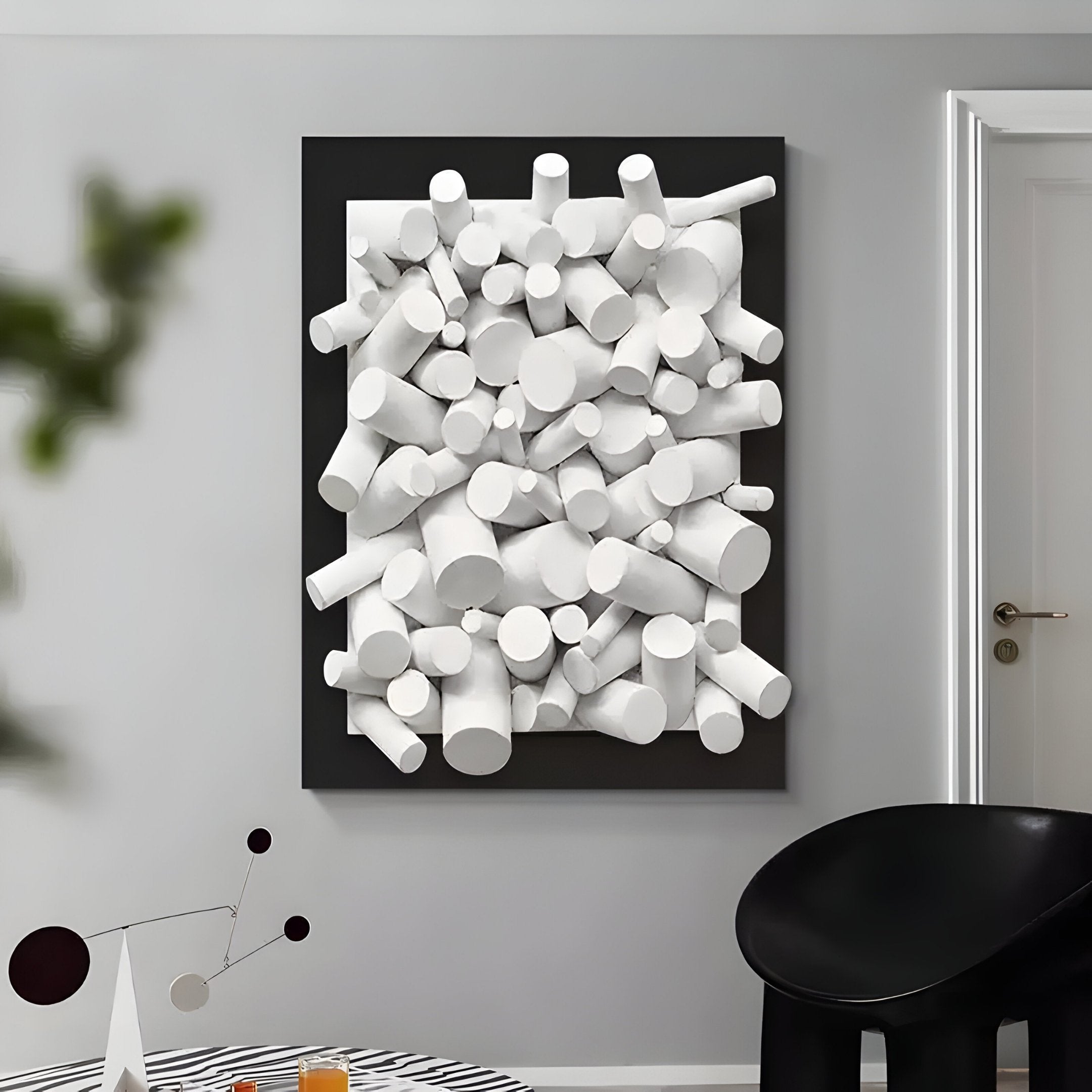 The Axion White Wood Cluster & Black Edge 3D Wall Art by Giant Sculptures features white cylindrical shapes in a black frame on a gray wall, complementing the rooms contemporary interiors with a black chair, patterned table, and small mobile sculpture.