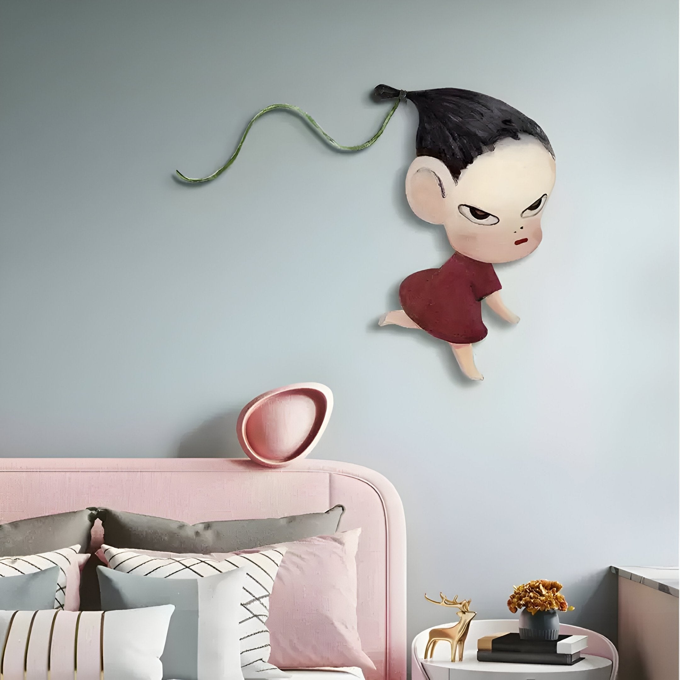 A minimalist bedroom includes a pale pink bed with striped pillows and a small table featuring a golden deer and potted plant. Above the bed, Giant Sculptures Enigmo Playful Cartoon Character Wooden Wall Art showcases a child with a long braided ponytail on a soft grayish-blue wall.