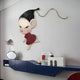 The Enigmo Playful Cartoon Character Wooden Wall Art by Giant Sculptures, showing a serious figure with a red outfit and ponytail, is mounted above a modern blue shelf. The shelf displays a vase with silver leaves and minimalistic decorative items.