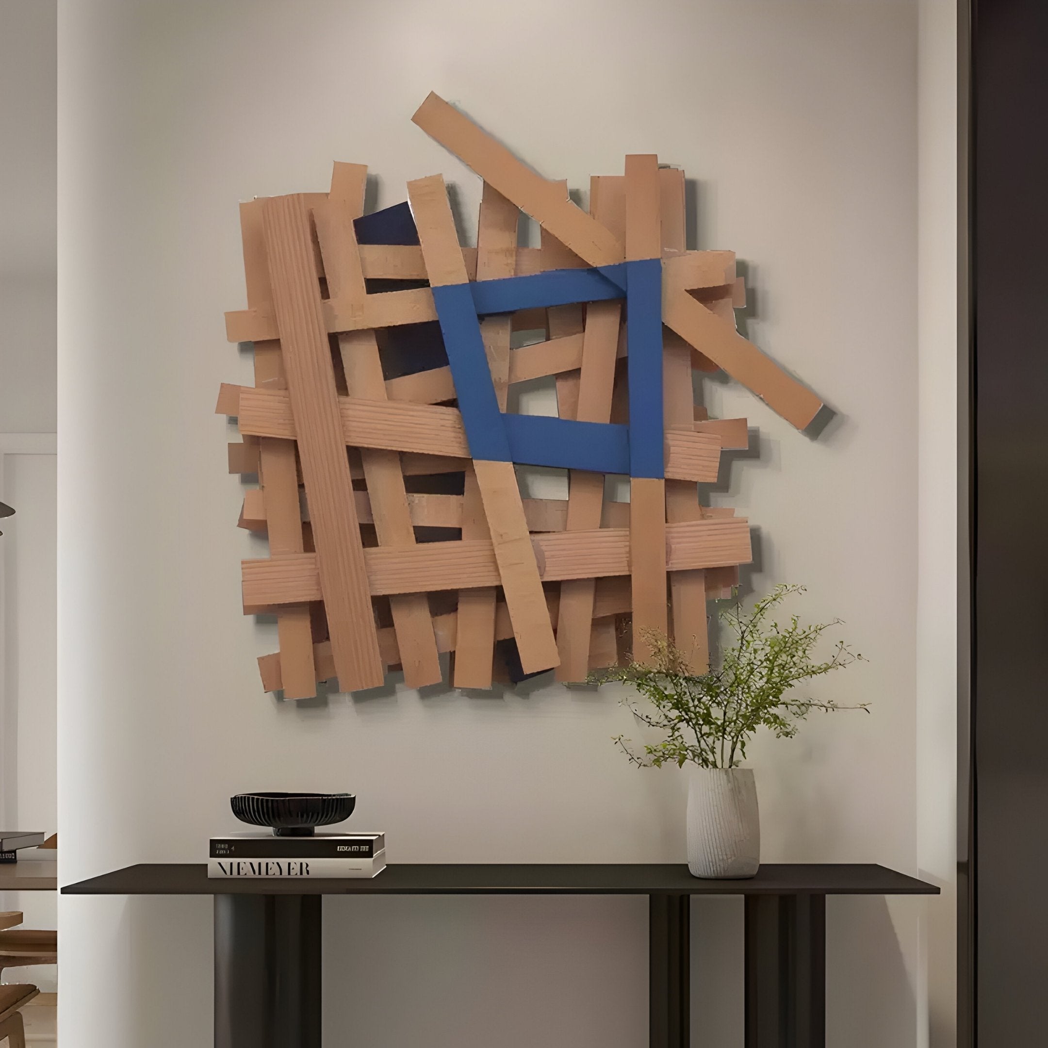 The Lignos Blue Accent Layered Wooden Strips 3D Wall Art from Giant Sculptures hangs on a white wall. Below, a minimalist table displays a book, small black dish, and vase with green foliage.