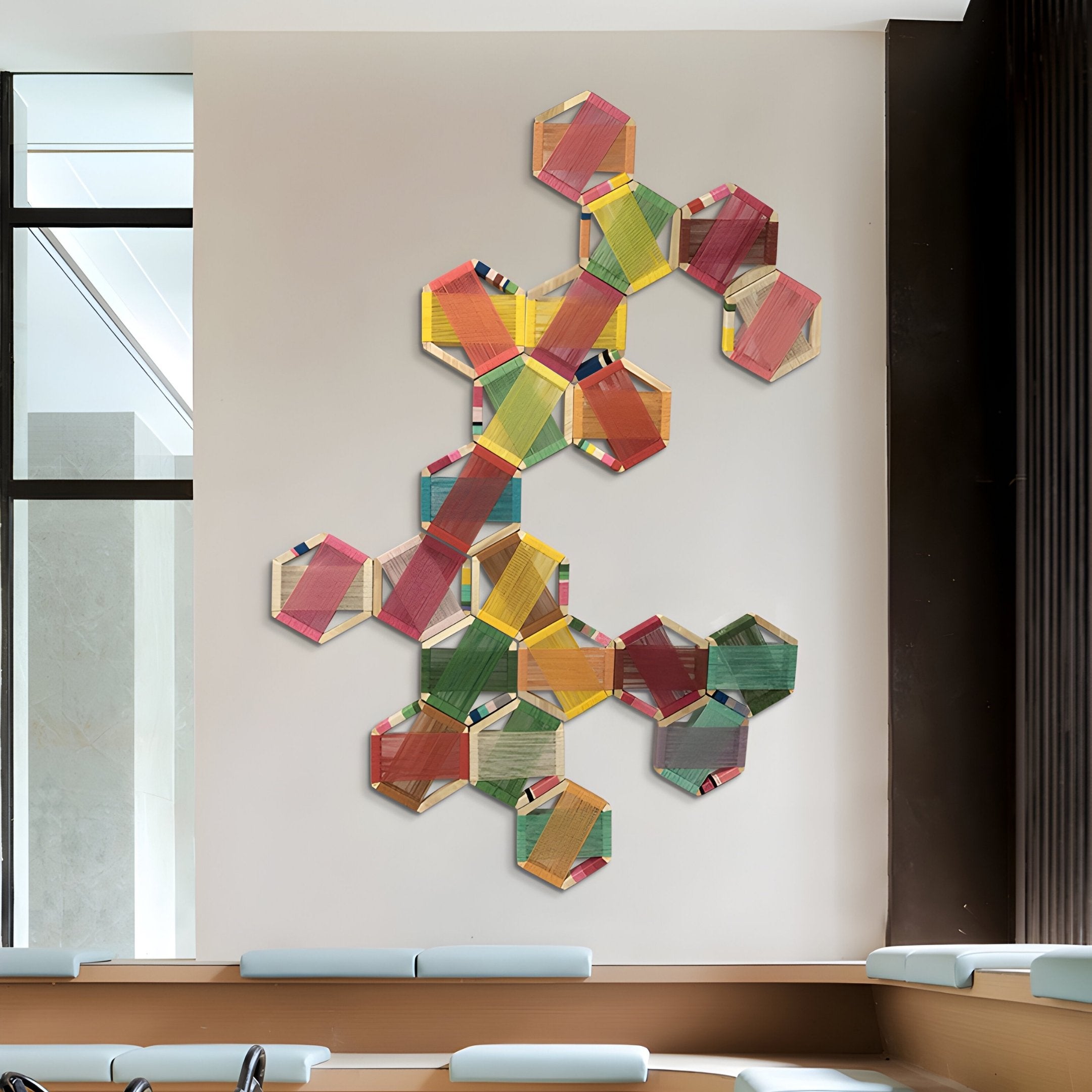 The Spectra V Multicoloured Interwoven Hexagonal Frame 3D Wall Art from Giant Sculptures features a geometric design of layered, striped hexagons. It stands out on a light backdrop by a large window, with inviting benches cushioned in light blue beneath it.