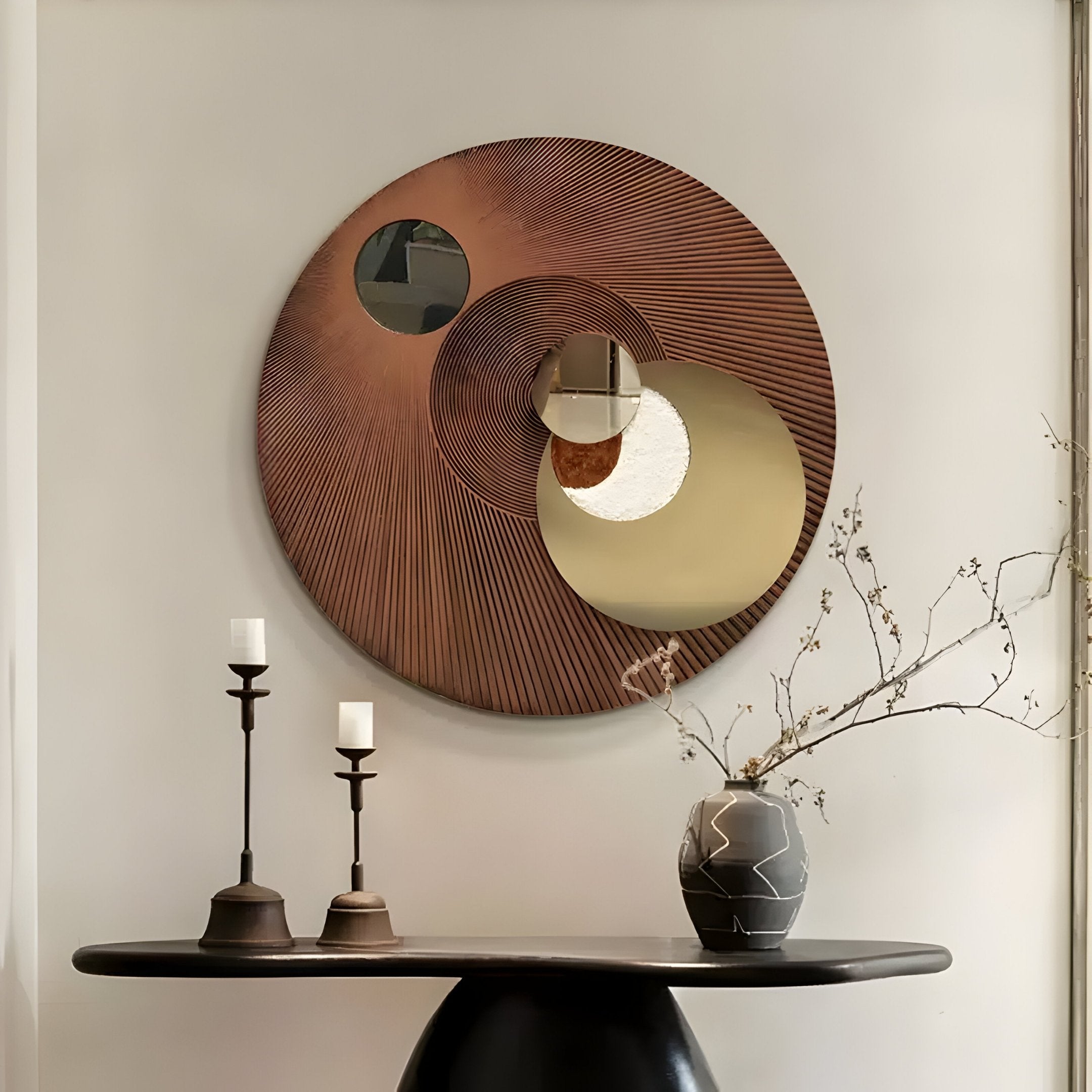 The wall showcases Giant Sculptures Orbitra Copper Reflective Circular 3D Wall Art. Below, a dark round table holds two candlesticks and a vase with bare branches, altogether creating a minimalist and modern aesthetic.