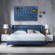 A modern bedroom features a blue upholstered bed with white bedding and geometric pillows, flanked by two white nightstands with gold-accented lamps. Above, the Orbison Blue 3D Wood Relief Wall Art With Floating Metal Ball from Giant Sculptures hangs alongside blue and gold abstract art. A light rug completes the look.