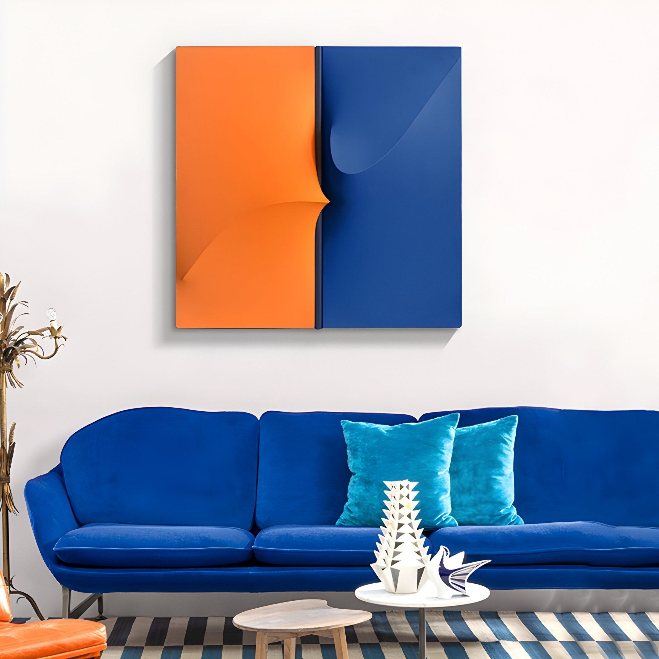 A modern living room has a blue velvet sofa with turquoise pillows and Delinea Dual Contrast Orange & Blue Fabric 3D Wood Relief Wall Art by Giant Sculptures above. A striped rug, decorative white sculptures, and a circular wooden table complete the look.