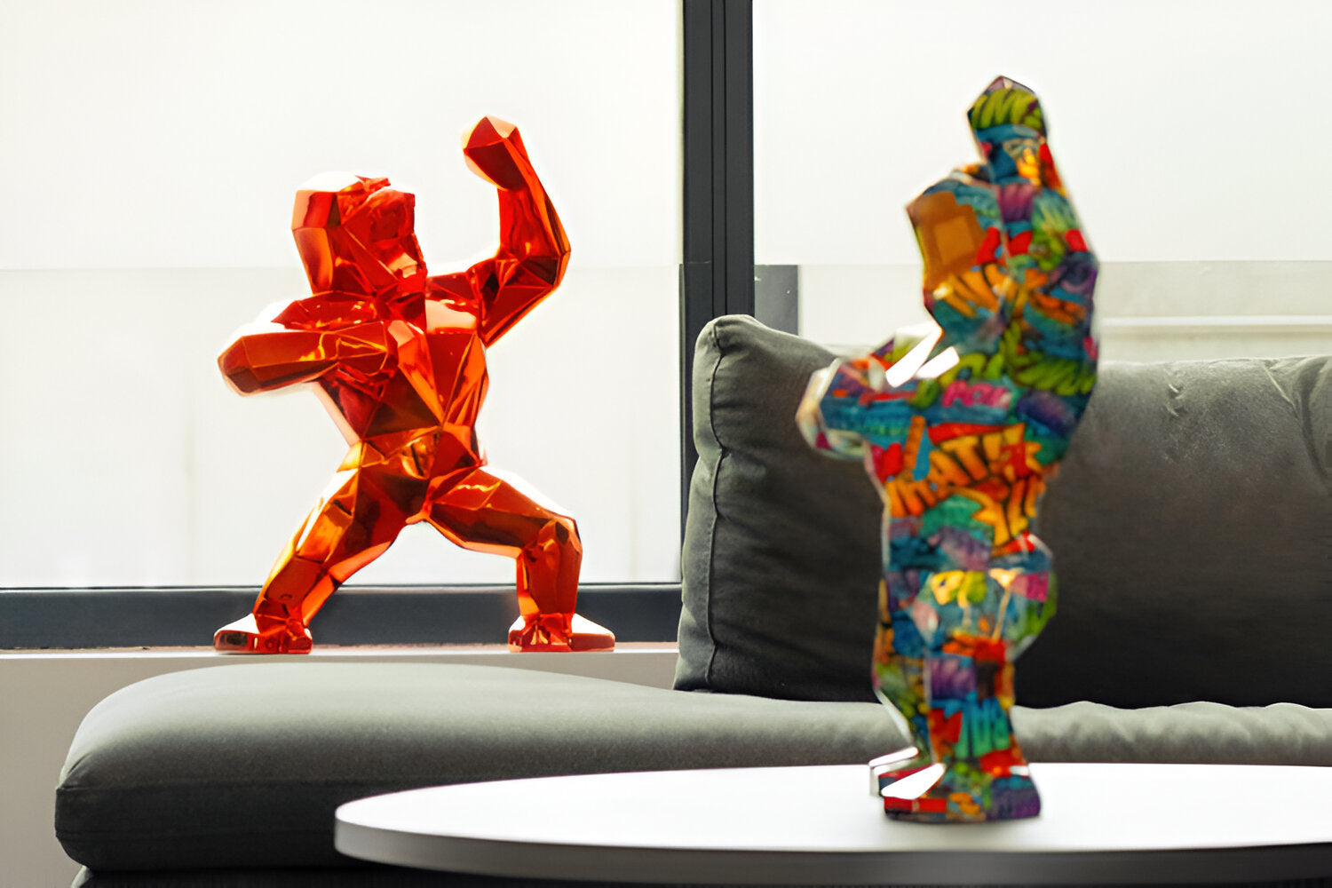 Two colorful gorilla sculptures stand in a modern living space. One is bright orange and positioned near a window, while the other features a vibrant, multicolored design and is placed on a table near a sofa.