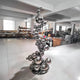 The Molecular Silver Organic Steel Outdoor Sculpture by Giant Sculptures, standing at 226cm, features interconnected shiny silver spheres of varying sizes. It is elegantly placed on a polished floor in a spacious studio with industrial-style windows and shelves in the background.