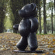 The Gacko Black LV Bear Sculpture by Giant Sculptures stands 165cm tall on a leaf-strewn path, surrounded by trees. Its embossed logos give it a luxurious look, evoking a contemporary park setting in autumn.