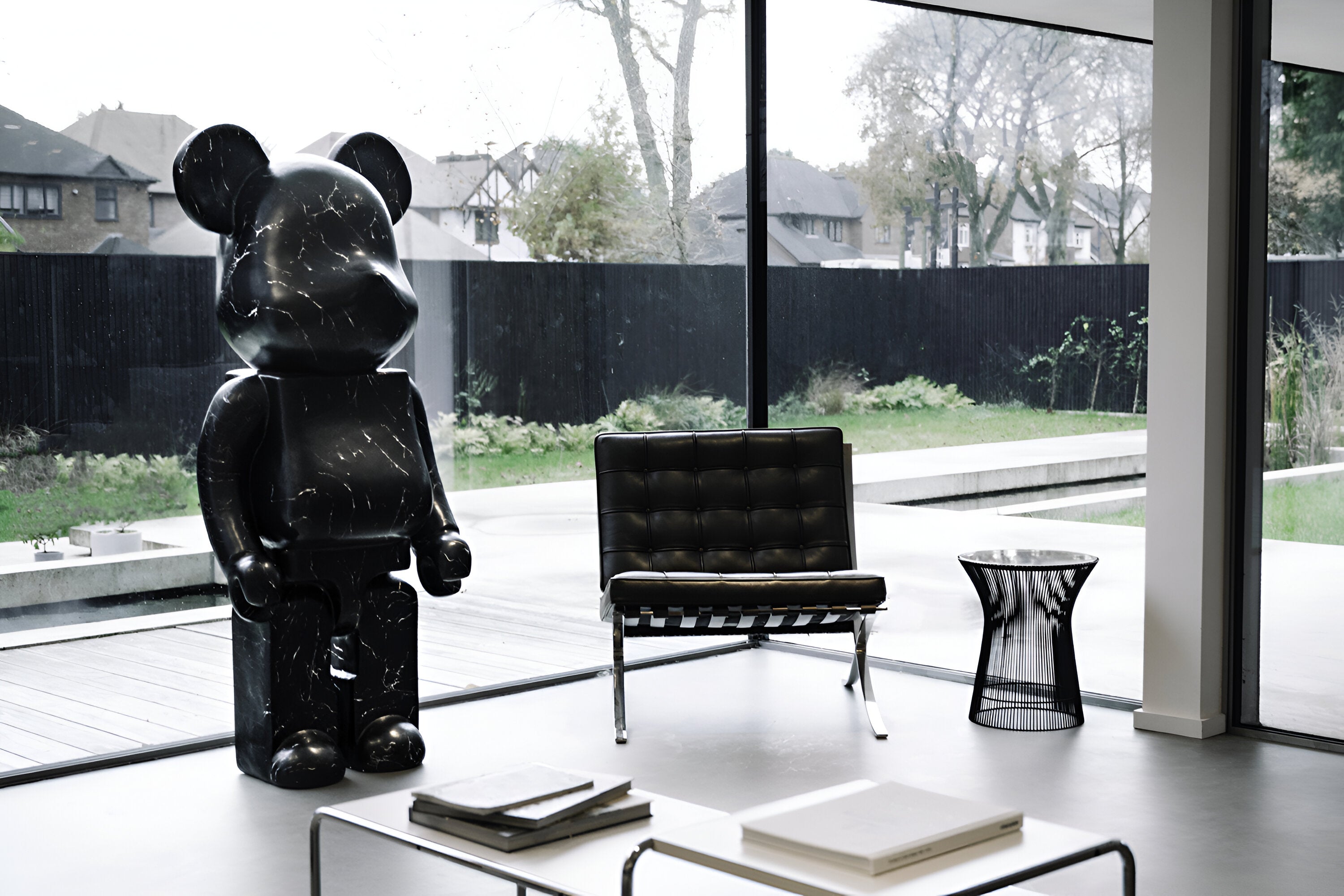 A tall, black marble-style bear sculpture standing in a modern living room with large floor-to-ceiling windows. The sculpture is placed next to a black leather chair and a side table, contrasting against the serene, outdoor garden visible through the glass.