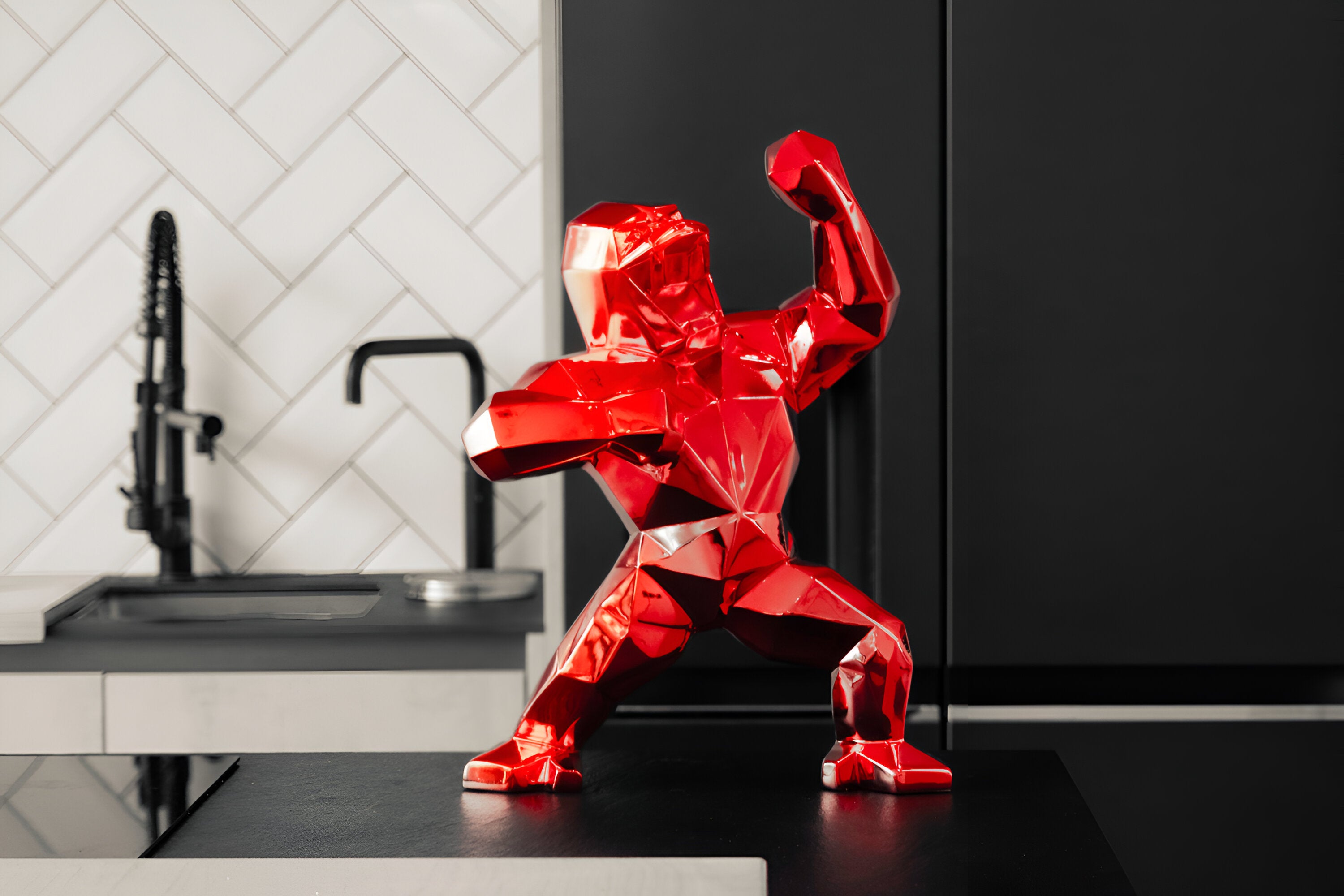 Vibrant red geometric gorilla sculpture with a glossy finish, placed on a black kitchen counter, contrasted with the sleek black kitchen fixtures and white herringbone tiled wall.