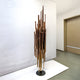 The Strata Copper Abstract Steel Sculpture by Giant Sculptures, standing at 165/220cm, features vertically arranged bronze rods of varying lengths on a round base. Its clustered, organic look with copper patina complements the minimalist room against a plain white wall.